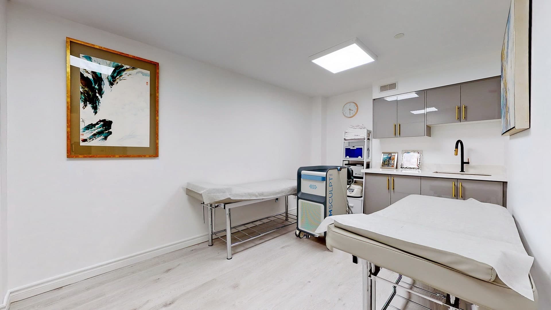 Bright medical treatment room featuring two examination beds, an Emsculpt machine, and gray cabinetry.